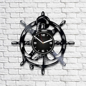 Puede incluir: Reloj de vinilo negro con temática náutica. El reloj presenta un timón de barco con un ancla en el centro. La esfera del reloj es negra con números y manecillas blancas.