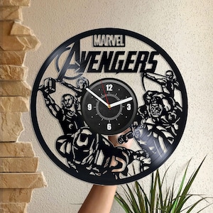 Pode incluir: Relógio de vinil preto com os personagens dos Vingadores da Marvel. O mostrador do relógio é preto com números e ponteiros brancos. O relógio é rodeado por um disco de vinil preto com um design recortado dos personagens dos Vingadores. O texto "MARVEL AVENGERS" está impresso no mostrador do relógio.