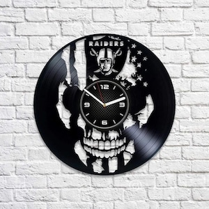 Puede incluir: Reloj de vinilo negro con un diseño de calavera y las palabras "RAIDERS" en blanco. La esfera del reloj es una calavera en blanco y negro con un diseño de bandera estadounidense blanca.