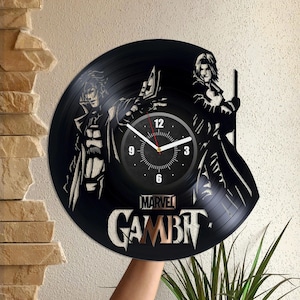 May include: Black vinyl record clock featuring a silhouette of two characters from the Marvel Comics series Gambit. The clock face is white with black numbers and hands. The text "MARVEL GAMBIT" is printed below the clock face.
