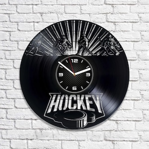 Puede incluir: Reloj de vinilo negro con temática de hockey. La esfera del reloj presenta una imagen en blanco y negro de jugadores de hockey y la palabra "HOCKEY" en letras blancas. Las manecillas del reloj son negras y blancas.