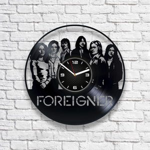 Könnte beinhalten: Eine schwarze Schallplattenuhr mit einer Silhouette der Band Foreigner. Das Zifferblatt der Uhr hat ein schwarz-weißes Gittermuster mit silbernen Zeigern und Zahlen.