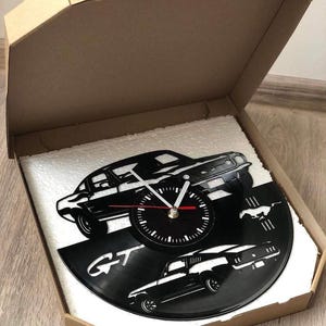 May include: A black vinyl record clock featuring a silhouette of a Ford Mustang GT. The clock face is black with white hands and numbers. The record is mounted on a white foam board inside a brown cardboard box.