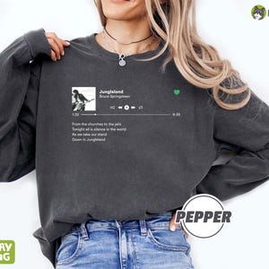 May include: A dark gray long-sleeve shirt with a white graphic of a music player screen displaying the song "Jungleland" by Bruce Springsteen. The lyrics "From the churches to the jails Tonight all is silence in the world As we take our stand Down in Jungleland" are visible on the screen.