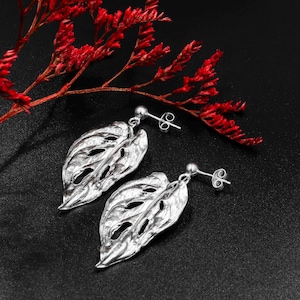 May include: Silver earrings shaped like a leaf with a cut-out design. The earrings are on a black background with red flowers.
