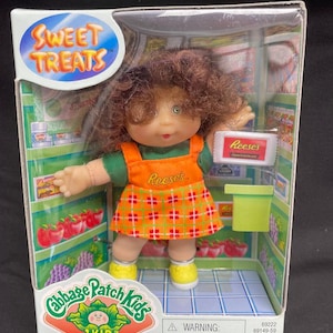 May include: A Cabbage Patch Kids doll dressed as a cashier in a grocery store. The doll is wearing a green shirt, an orange and red plaid skirt, and yellow shoes. The doll is holding a box of Reese's peanut butter cups. The doll is standing in front of a toy grocery store with shelves stocked with toy food items. The box has the words "Sweet Treats" and "Cabbage Patch Kids" on it. The box also has a warning label that says "Choking Hazard - Small parts. Not for children under 3 years. For ages over 3 years."