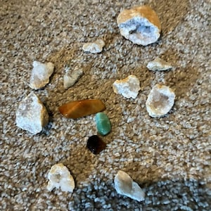 May include: A collection of various small stones and crystals, including several white geode fragments with crystalline interiors, one smooth amber-colored stone, one small polished green stone, and one dark brown polished stone.