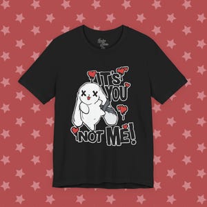 May include: A black short-sleeve t-shirt featuring a white graphic of a cartoon rabbit with X-shaped eyes and a tongue sticking out. The text reads IT'S YOU NOT ME! in white block letters, surrounded by several small red dripping heart illustrations.