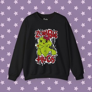 May include: A black long-sleeve sweatshirt featuring a graphic print of a green zombie rabbit with X-shaped eyes and an exposed brain. The text ZOMBIE HUGS is printed in red with a white drip effect outline, centered on the chest of the garment.