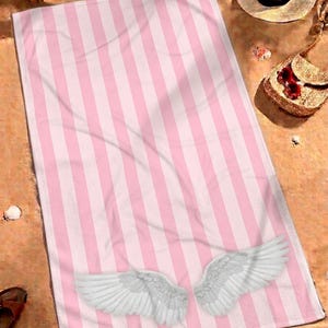 May include: A rectangular beach towel featuring a repeating vertical striped pattern in light pink and white. The lower portion of the towel displays a graphic of two large, detailed white feathered wings spread outward toward the bottom corners.