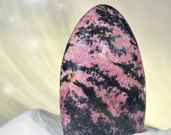 May include: A polished, oval-shaped rhodonite stone with a striking pattern of pink and black. The stone is set against a backdrop of white sand and a soft, yellow-toned background.