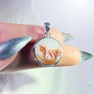May include: A round pendant with a silver-colored bail. The pendant features a white background with a natural, branching pattern in shades of orange and brown. The pendant is held between fingers.
