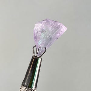 Op de afbeelding: Een ruwe, lichtpaarse edelsteen, vastgehouden door een zilveren metalen gereedschap. De edelsteen heeft een kristalstructuur met iriserende flitsen. Het gereedschap heeft een getextureerde handgreep.
