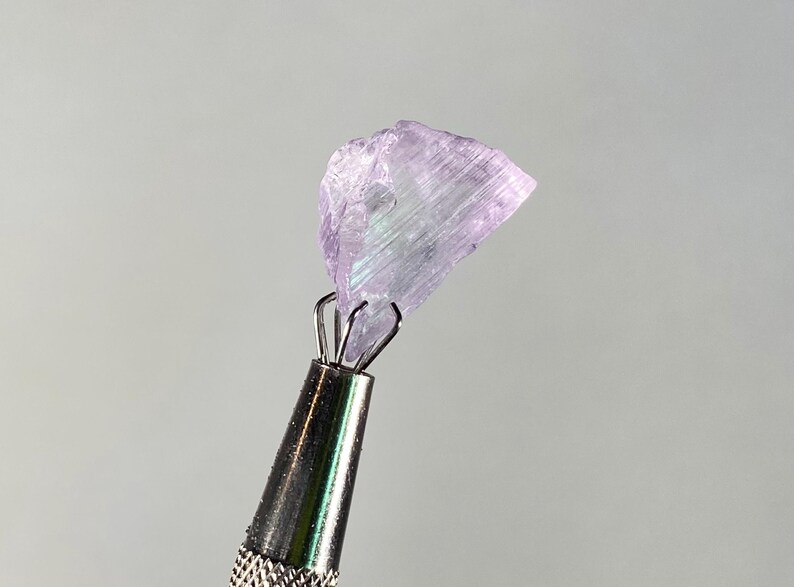 Op de afbeelding: Een ruwe, lichtpaarse edelsteen, vastgehouden door een zilveren metalen gereedschap. De edelsteen heeft een kristalstructuur met iriserende flitsen. Het gereedschap heeft een getextureerde handgreep.