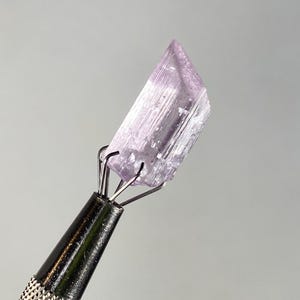 Op de afbeelding: Een lichtpaars kristal dat wordt vastgehouden door een metalen gereedschap. Het kristal is een rechthoekig prisma met zichtbare strepen en een licht schuine bovenkant. Het gereedschap heeft een zwarte, getextureerde handgreep en zilveren pinnen.