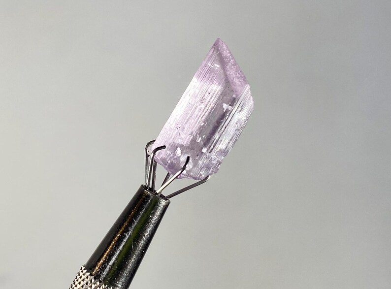 Op de afbeelding: Een lichtpaars kristal dat wordt vastgehouden door een metalen gereedschap. Het kristal is een rechthoekig prisma met zichtbare strepen en een licht schuine bovenkant. Het gereedschap heeft een zwarte, getextureerde handgreep en zilveren pinnen.