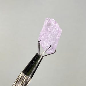 Op de afbeelding: Een lichtpaars, ruw kristal dat wordt vastgehouden door een metalen gereedschap met een zwarte, getextureerde handgreep. Het kristal heeft een ruw, ongelijkmatig oppervlak en wordt verlicht tegen een neutrale grijze achtergrond. De pinnen van het gereedschap grijpen het kristal stevig vast.