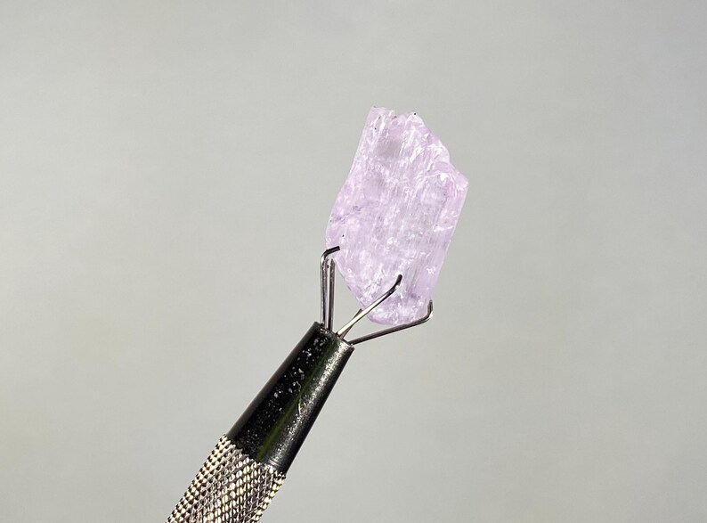 Op de afbeelding: Een lichtpaars, ruw kristal dat wordt vastgehouden door een metalen gereedschap met een zwarte, getextureerde handgreep. Het kristal heeft een ruw, ongelijkmatig oppervlak en wordt verlicht tegen een neutrale grijze achtergrond. De pinnen van het gereedschap grijpen het kristal stevig vast.