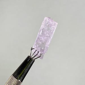 Op de afbeelding: Een lichtpaars kristal dat wordt vastgehouden door een zilveren metalen gereedschap. Het kristal is rechthoekig met een ruw, getextureerd oppervlak. Het gereedschap heeft een getextureerde handgreep en een klauwachtige grip die het kristal vasthoudt. De achtergrond is effen lichtgrijs.