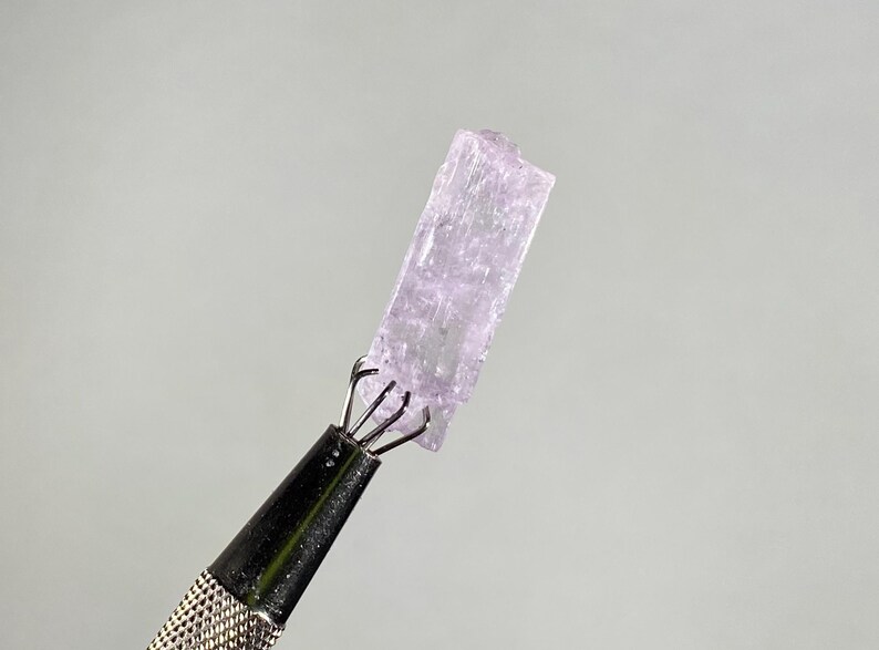 Op de afbeelding: Een lichtpaars kristal dat wordt vastgehouden door een zilveren metalen gereedschap. Het kristal is rechthoekig met een ruw, getextureerd oppervlak. Het gereedschap heeft een getextureerde handgreep en een klauwachtige grip die het kristal vasthoudt. De achtergrond is effen lichtgrijs.