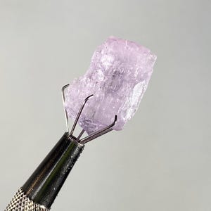 Op de afbeelding: Een ruwe, lichtpaarse edelsteen wordt vastgehouden door een metalen gereedschap met een getextureerde handgreep. De kristalsteen wordt verlicht tegen een lichtgrijze achtergrond. De tanden van het gereedschap grijpen de steen stevig vast.