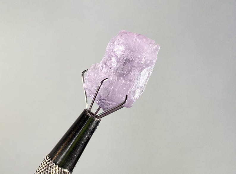 Op de afbeelding: Een ruwe, lichtpaarse edelsteen wordt vastgehouden door een metalen gereedschap met een getextureerde handgreep. De kristalsteen wordt verlicht tegen een lichtgrijze achtergrond. De tanden van het gereedschap grijpen de steen stevig vast.