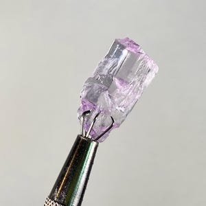 Op de afbeelding: Een ruw, kristallijn mineraalmonster, vastgehouden door een metalen gereedschap. Het mineraal heeft een doorschijnende, lichtpaarse kleur met zichtbare interne structuren. Het gereedschap heeft een zilveren metalen handvat.