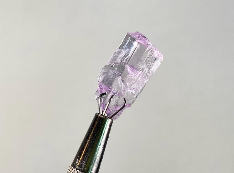 Op de afbeelding: Een ruw, kristallijn mineraalmonster, vastgehouden door een metalen gereedschap. Het mineraal heeft een doorschijnende, lichtpaarse kleur met zichtbare interne structuren. Het gereedschap heeft een zilveren metalen handvat.