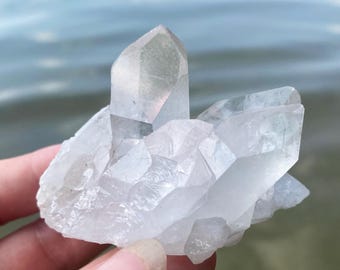 Op de afbeelding: Een cluster van heldere kwartskristallen met meerdere punten. De kristallen worden voor een vage waterachtergrond gehouden. Het kwarts heeft een doorschijnend uiterlijk en een natuurlijke, ruwe textuur.