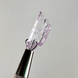 Op de afbeelding: Een kleine, onregelmatig gevormde, doorschijnende, lichtpaarse kristal wordt vastgehouden door een metalen gereedschap. De kristal heeft een gebroken uiterlijk met scherpe randen. Het gereedschap heeft een zilveren handvat en zwarte punt, tegen een lichtgrijze achtergrond.