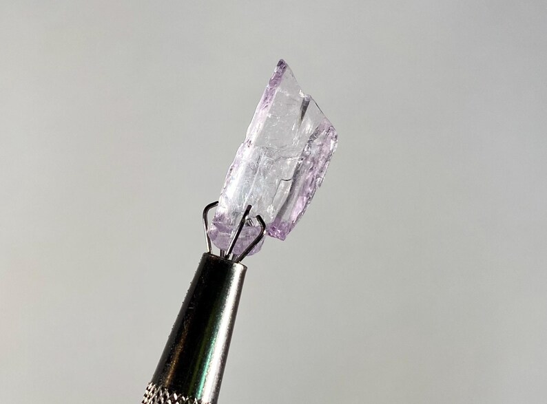 Op de afbeelding: Een kleine, onregelmatig gevormde, doorschijnende, lichtpaarse kristal wordt vastgehouden door een metalen gereedschap. De kristal heeft een gebroken uiterlijk met scherpe randen. Het gereedschap heeft een zilveren handvat en zwarte punt, tegen een lichtgrijze achtergrond.