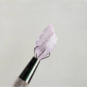 Op de afbeelding: Een lichtpaars kristal vastgehouden door een metalen gereedschap. Het kristal heeft een gelaagde, getextureerde uitstraling met een lichtroze tint aan de bovenkant. De tool heeft een getextureerde handgreep en een zilveren afwerking.