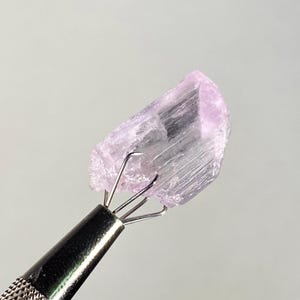 Op de afbeelding: Een lichtpaarse, ruwe edelsteen die wordt vastgehouden door een metalen gereedschap. De edelsteen heeft een kristalstructuur en is licht lavendel van kleur. Het gereedschap heeft een getextureerde handgreep en vier pinnen die de steen vasthouden.