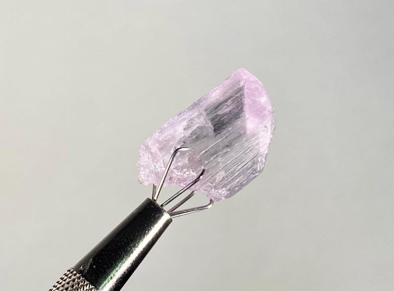 Op de afbeelding: Een lichtpaarse, ruwe edelsteen die wordt vastgehouden door een metalen gereedschap. De edelsteen heeft een kristalstructuur en is licht lavendel van kleur. Het gereedschap heeft een getextureerde handgreep en vier pinnen die de steen vasthouden.