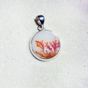 May include: A round pendant with a silver bail. The pendant features a white background with a natural, tree-like pattern in shades of red and orange. The pendant is set in a silver bezel.