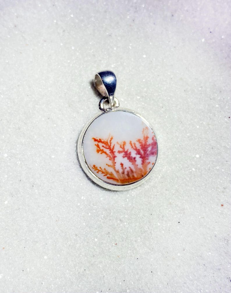 May include: A round pendant with a silver bail. The pendant features a white background with a natural, tree-like pattern in shades of red and orange. The pendant is set in a silver bezel.
