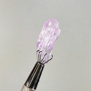 Op de afbeelding: Een lichtpaars kristal dat wordt vastgehouden door een metalen gereedschap. Het kristal heeft een ruw, getextureerd oppervlak en een doorschijnend uiterlijk. Het gereedschap heeft een zilveren handvat en meerdere pinnen om het kristal vast te houden. De achtergrond is effen lichtgrijs.