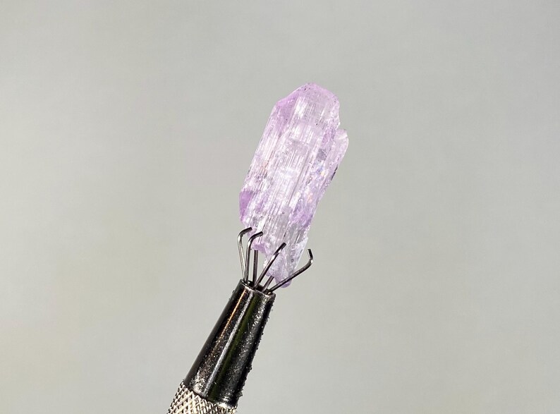 Op de afbeelding: Een lichtpaars kristal dat wordt vastgehouden door een metalen gereedschap. Het kristal heeft een ruw, getextureerd oppervlak en een doorschijnend uiterlijk. Het gereedschap heeft een zilveren handvat en meerdere pinnen om het kristal vast te houden. De achtergrond is effen lichtgrijs.