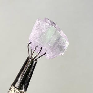 Op de afbeelding: Een lichtpaarse, ruwe edelsteen die wordt vastgehouden door een metalen gereedschap. De edelsteen heeft een kristallijne structuur met een doorschijnende kwaliteit. Het gereedschap heeft een getextureerde handgreep en meerdere pinnen om de steen vast te zetten. De achtergrond is effen grijs.