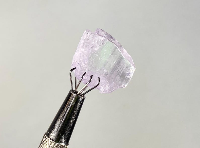 Op de afbeelding: Een lichtpaarse, ruwe edelsteen die wordt vastgehouden door een metalen gereedschap. De edelsteen heeft een kristallijne structuur met een doorschijnende kwaliteit. Het gereedschap heeft een getextureerde handgreep en meerdere pinnen om de steen vast te zetten. De achtergrond is effen grijs.