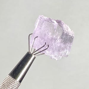 Op de afbeelding: Een lichtpaars, kristallijn mineraal specimen wordt vastgehouden door een metalen klauwgereedschap. Het mineraal heeft een ruw, getextureerd oppervlak en een doorschijnend uiterlijk. De tool heeft een getextureerde handgreep.