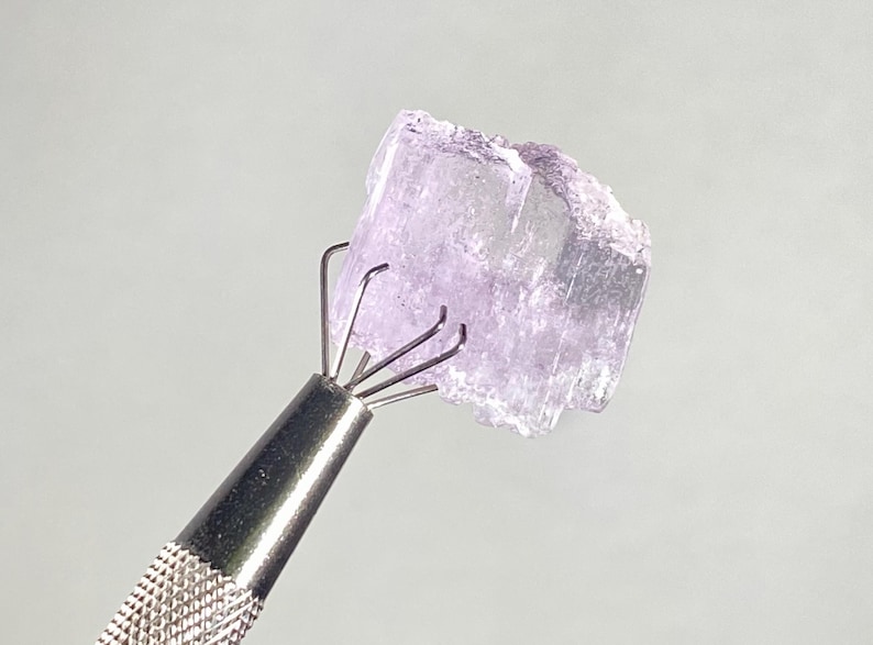 Op de afbeelding: Een lichtpaars, kristallijn mineraal specimen wordt vastgehouden door een metalen klauwgereedschap. Het mineraal heeft een ruw, getextureerd oppervlak en een doorschijnend uiterlijk. De tool heeft een getextureerde handgreep.