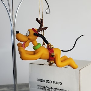May include: A yellow Pluto Christmas ornament with brown antlers, a red collar with gold bells, and a green collar. The ornament has a black tail and is suspended from a gold string. The ornament is on top of a white box.