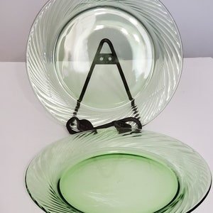 May include: Two round, light green glass plates featuring a swirling, spiral pattern radiating from the centre to the rim. One plate is positioned upright on a black metal display stand, while the second plate rests horizontally in the foreground.