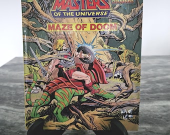 Puede incluir: Un cómic vintage de "Masters of the Universe" titulado "Maze of Doom". La portada presenta una ilustración dinámica de He-Man montado en una criatura verde y naranja, luchando en un entorno forestal. El cómic está sostenido por un soporte negro.