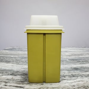 May include: A vintage olive green rectangular container with a white lid. The container has a ridged design on the sides and a slightly flared top. The lid is a contrasting white color and has a stepped design. The container is likely made of plastic.