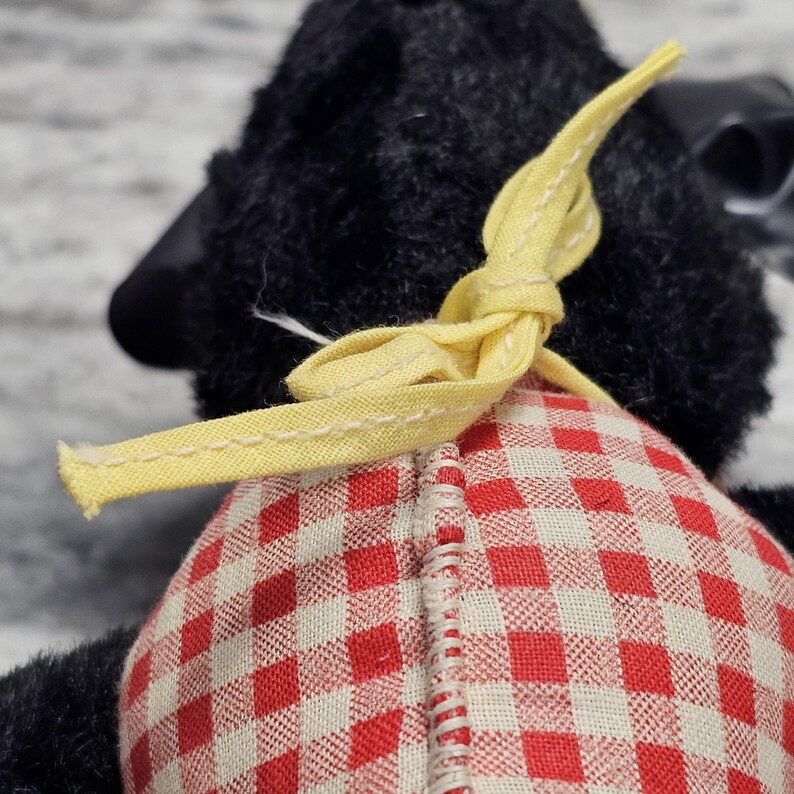 May include: A black plush toy sheep with a red and white checkered pattern on its back. The sheep has a yellow ribbon tied around its neck. The toy is made of soft, plush material.