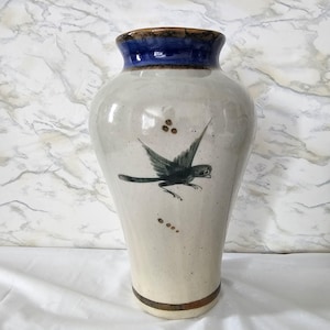 May include: A tall, off-white ceramic vase with a blue rim and a brown base. The vase features a hand-painted design of a dark teal bird in flight. The vase is approximately 30 cm tall.