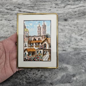 May include: A framed watercolor painting of a European townscape with a church and buildings with orange tile roofs. The painting is in a gold-colored frame and has the text "Axio 66 Venice S" in the lower right corner.