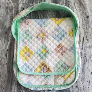 Peut inclure: Un sac en tissu matelassé avec un motif en losanges jaune pastel, bleu et rose. Le tissu présente des personnages Cabbage Patch Kids et des ballons. Le sac a une bordure vert menthe et un rabat avec le texte 1983 OAA, INC.